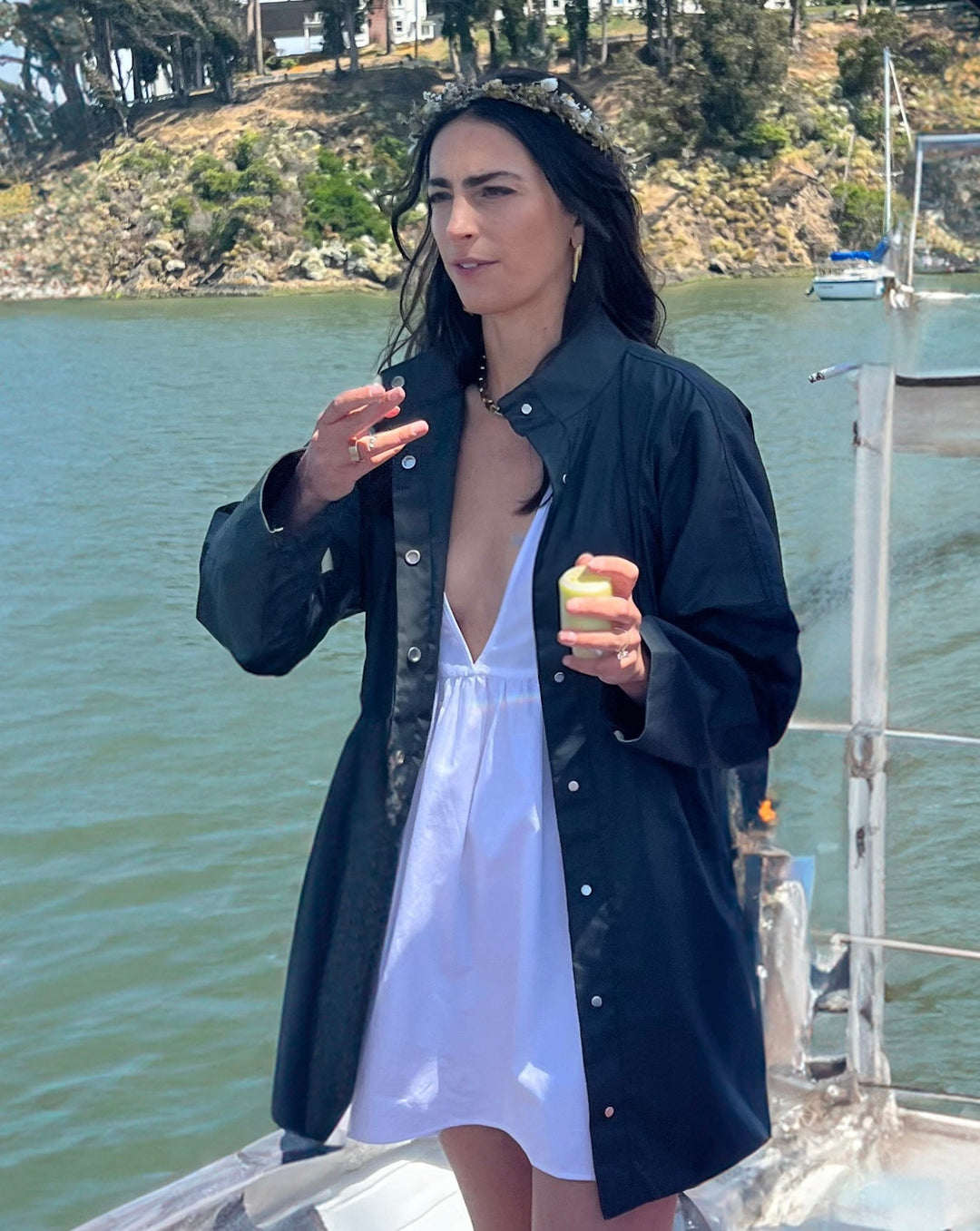 Model on a boat, wearing a stylish open black coat over a white dress, holding a drink in her hand. The background features a view of a bay with hills and other boats.