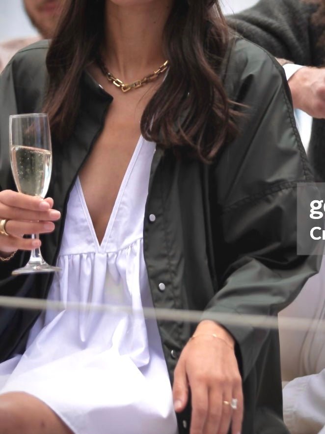 Close-up photo of a woman holding a champagne glass, dressed in a white V-neck dress under a dark grey coat.