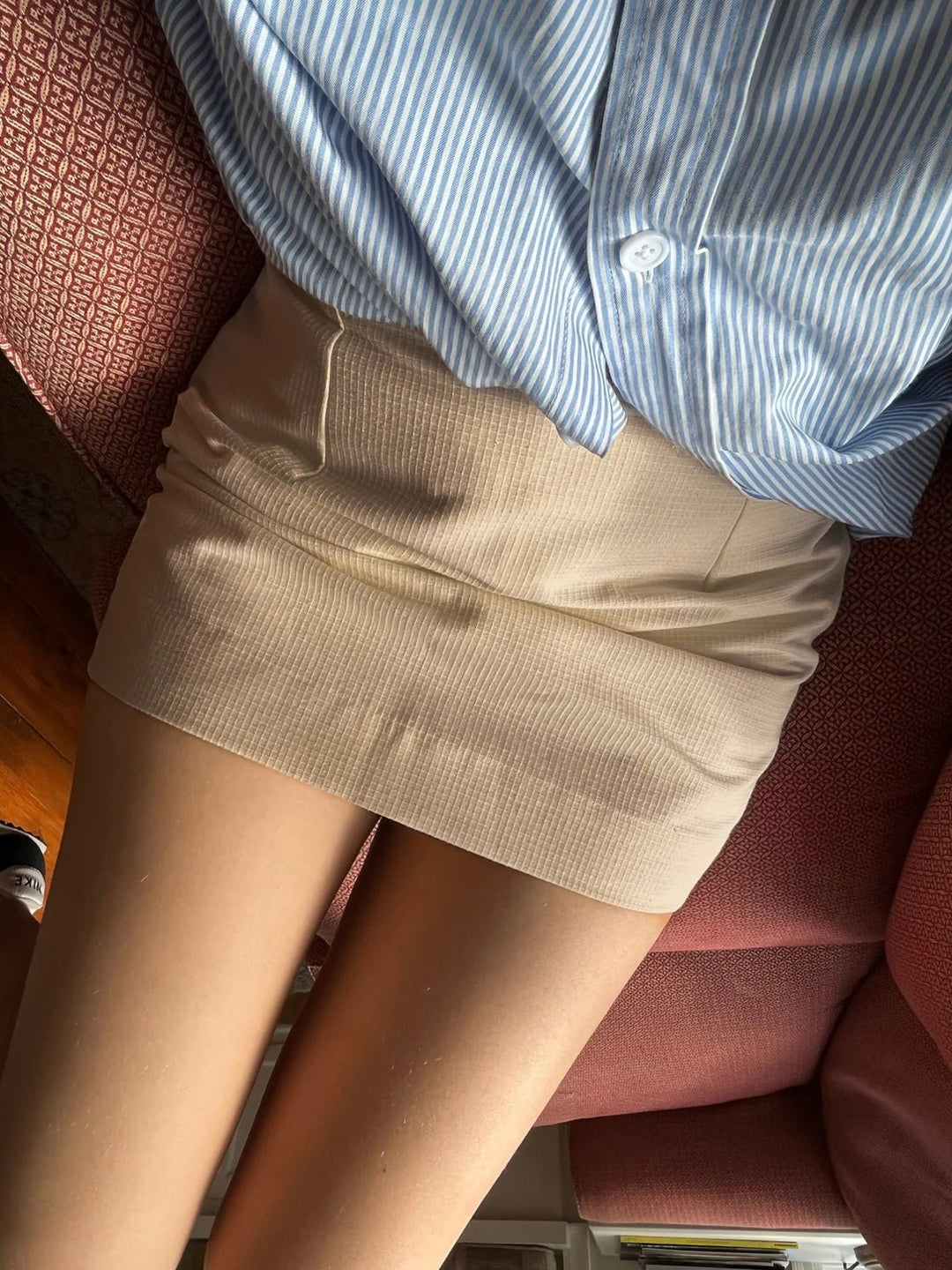 Model wearing a beige mini skirt paired with a blue striped button-up shirt, seated on a red couch. 