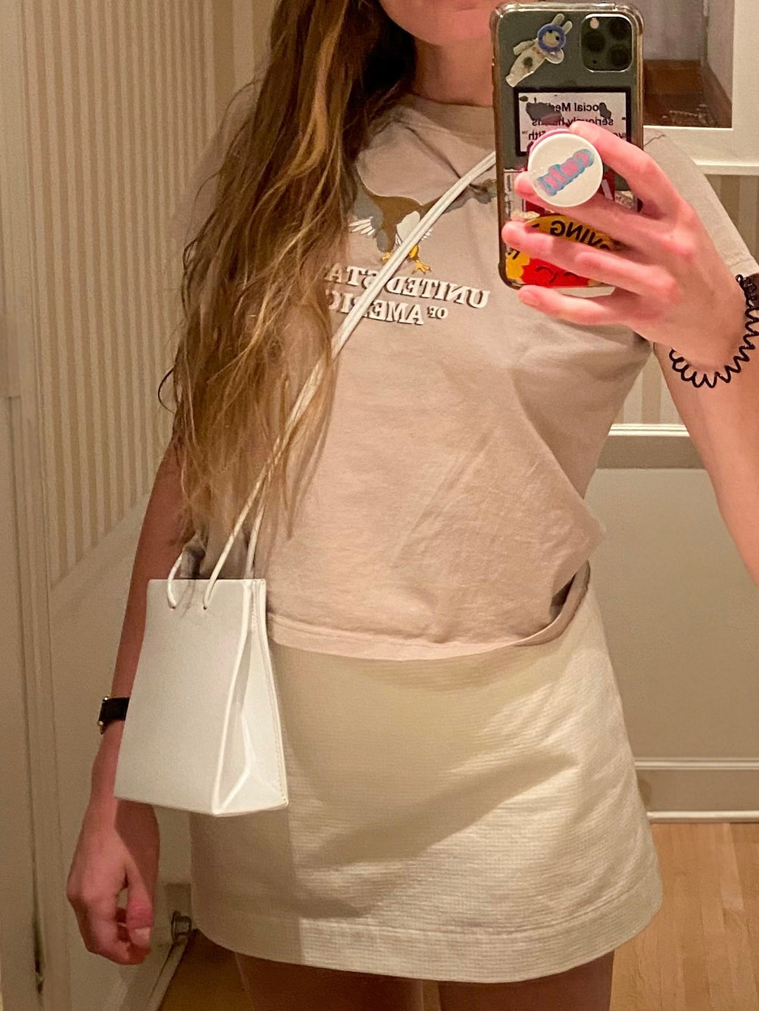 Selfie of a model in a casual outfit featuring a beige graphic t-shirt tucked into a white mini skirt. Accessorized with a small white shoulder bag and takes the photo in a mirror.