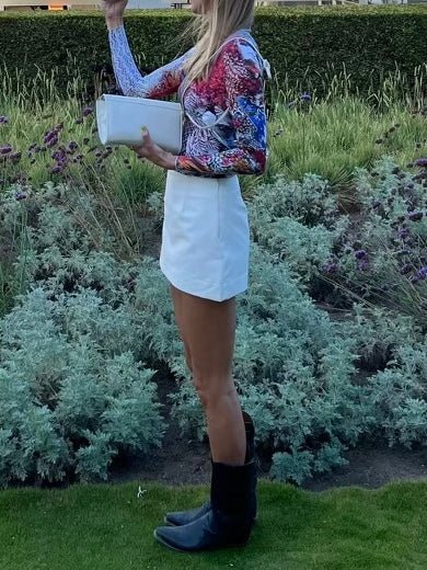 Photo of a woman in a colorful blouse and white skirt, wearing black boots and holding a white bag, standing in a garden with purple and green plants.