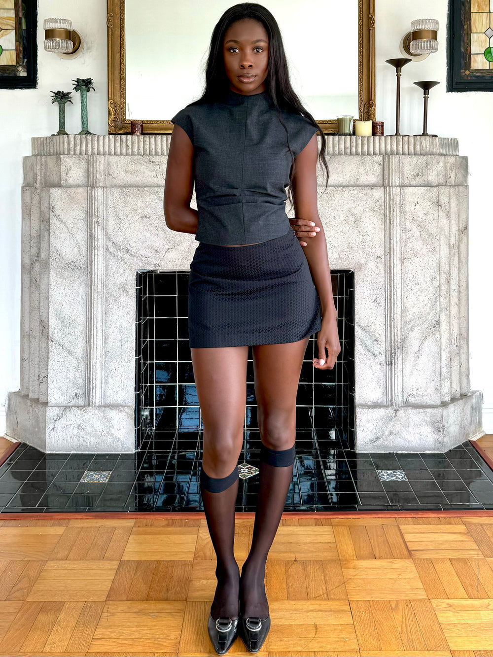 Model wearing a black textured mini skirt, fitted charcoal top, sheer knee-high socks, and black loafers with metal buckles, standing in front of a decorative fireplace.