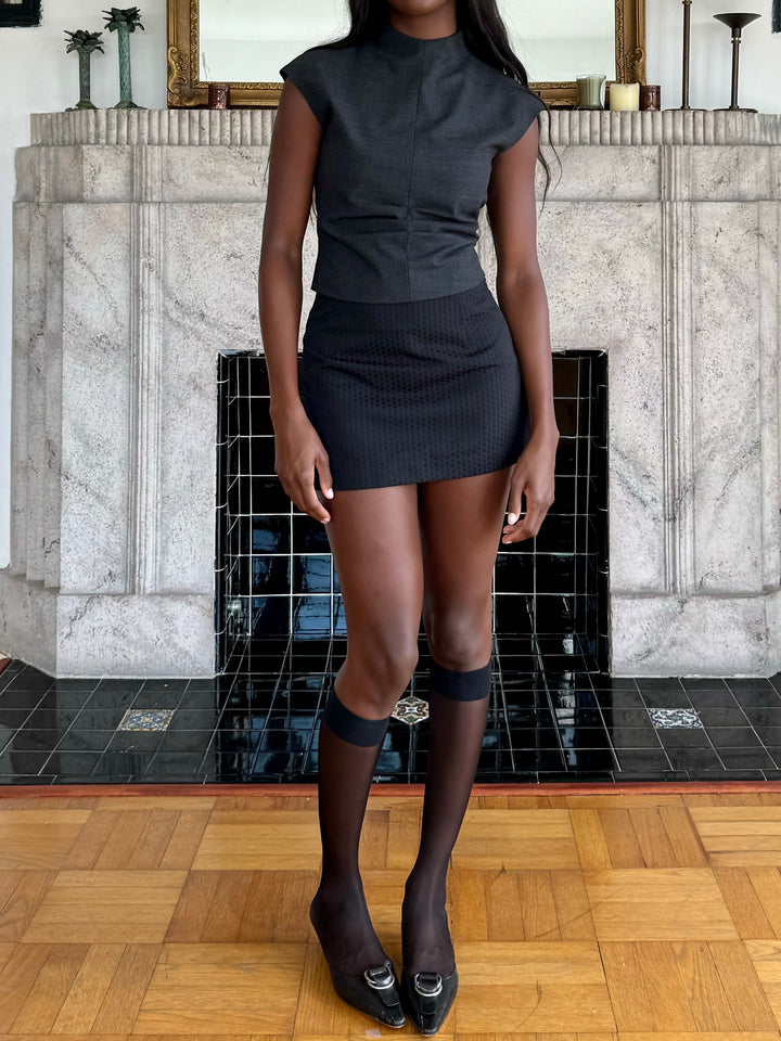 Model wearing a fitted charcoal top, black textured mini skirt, sheer knee-high socks, and black loafers with metal buckles, posing in front of a decorative fireplace.