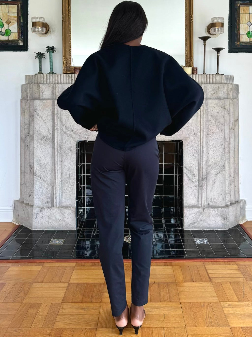 Model facing away from the camera, wearing an oversized black sweater with structured sleeves, black pants, and black heels, posing in front of a decorative fireplace.