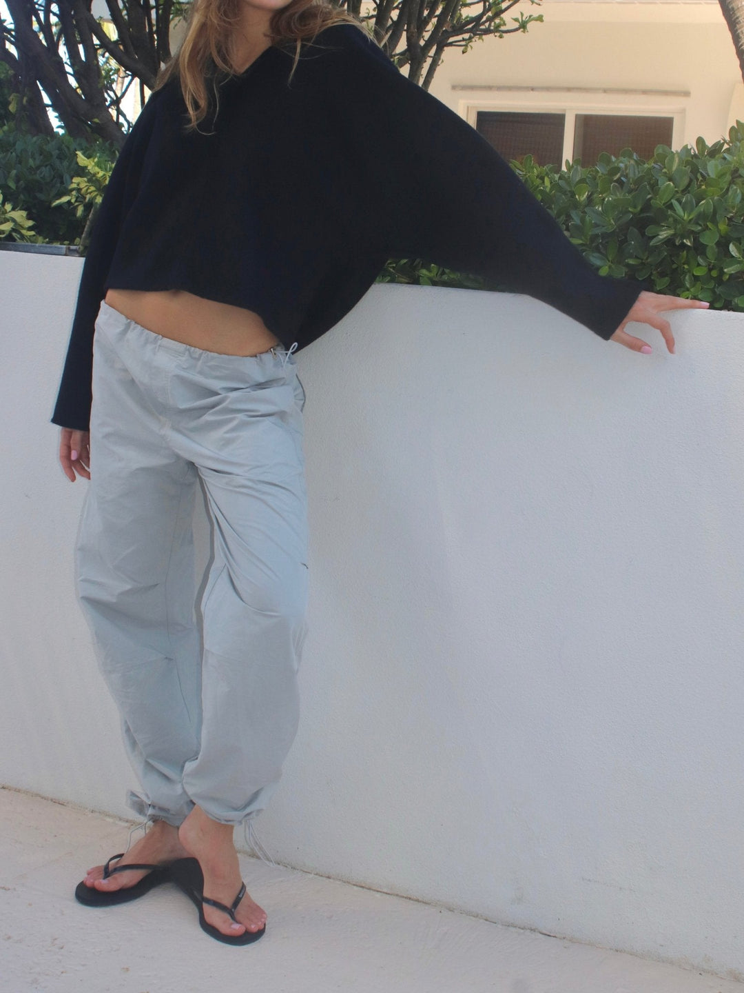 Model posing in a relaxed stance wearing a cropped black sweater and light gray pants, leaning against a white wall outdoors.