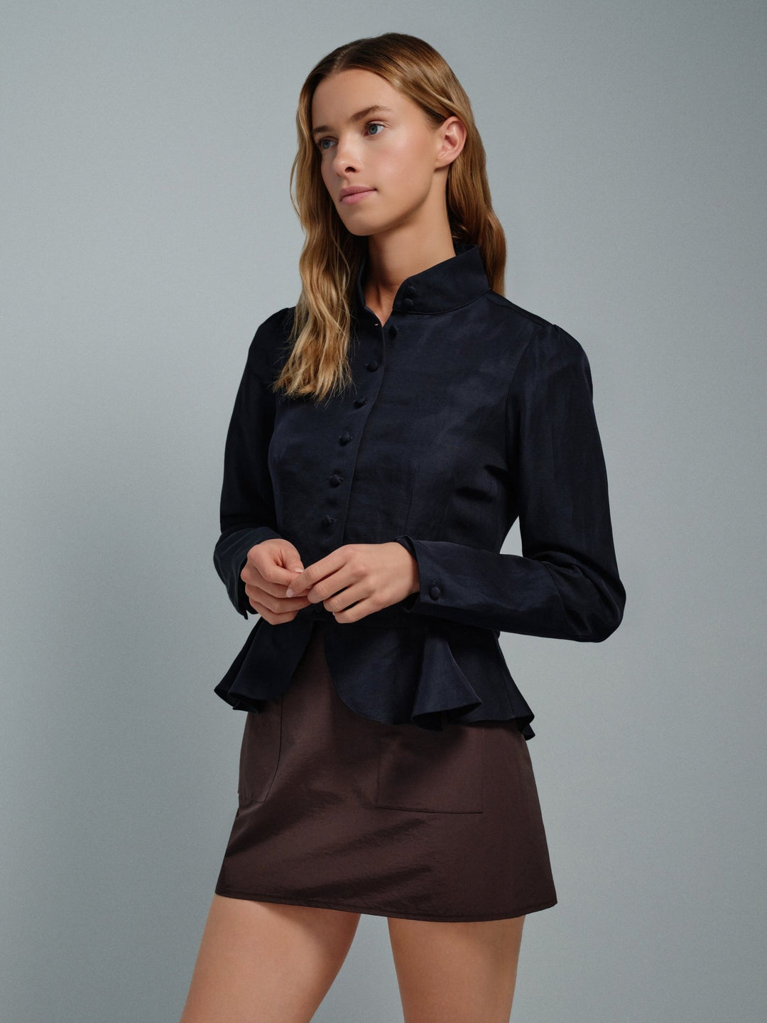Woman wearing a navy high collar jacket blouse and brown skirt against a gray background