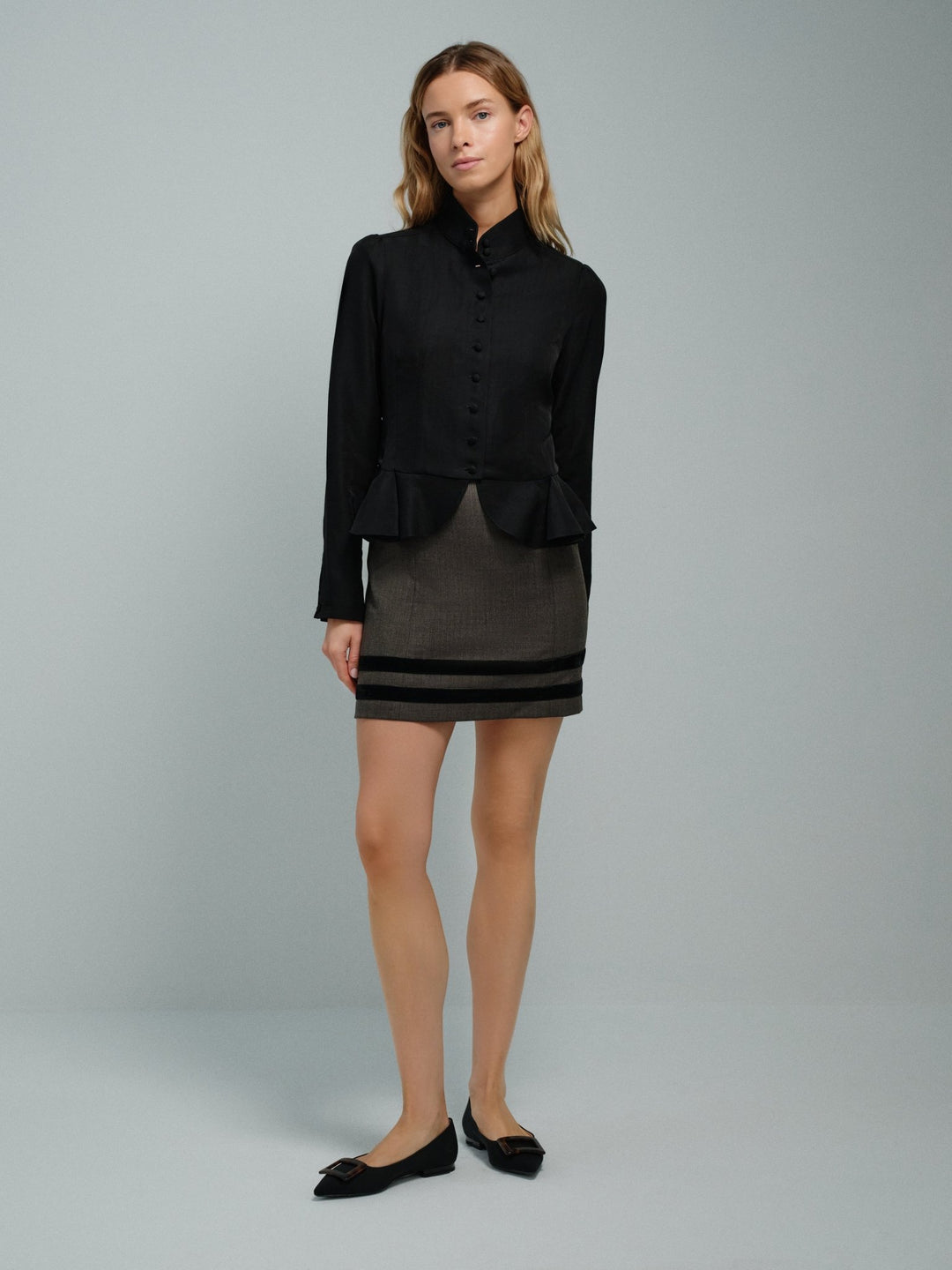 Woman wearing a black jacket and gray skirt with black accents on a gray background