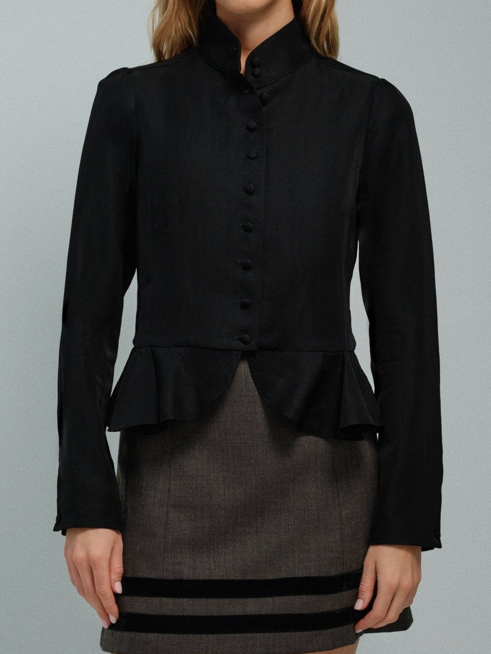 Woman wearing a black high collar jacket with a peplum waist over a gray dress on a gray background